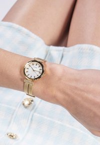 Montre-bracelet en maille dorée avec un cadran blanc, un boîtier rond, des index en or et une boucle décorative. Portée sur un poignet à la peau claire.