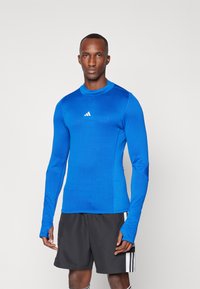 Blå atletisk langermet skjorte med rund krage og hvit logo på brystet, laget av et glatt, elastisk materiale. Kombinert med svarte shorts.