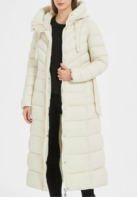 Manteau matelassé long en ivoire avec capuche. Comprend une fermeture éclair à l'avant et des boutons-pression, des poches latérales et des coutures horizontales pour ajouter de la texture.