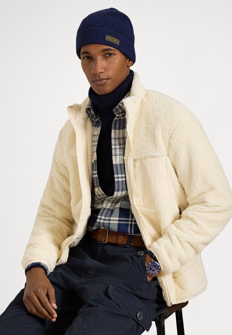 Άντρας που φοράει μπλε σκουφί, κρεμ μπουφάν από φ�ανέλα, μπλε πουκάμισο με καρό σχέδιο, σκούρο παντελόνι και ρολόι χειρός, καθισμένος μπροστά σε απλό λευκό φόντο.