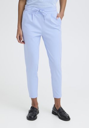 Person iført lyseblå taperede bukser med snøre i taljen og sorte vævede læderloafers, stående med den ene hånd i lommen.