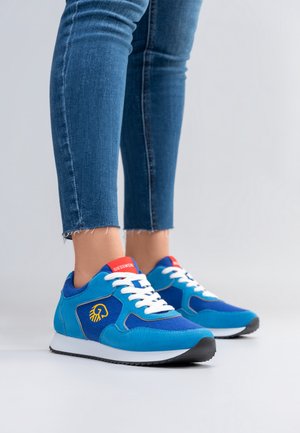 Osoba nosząca niebiesko-turkusowe trampki z białymi sznurowadłami i czerwonymi akcentami, stojąca na białej podłodze, ubrana w niebieskie dżinsy z postrzępionymi brzegami.