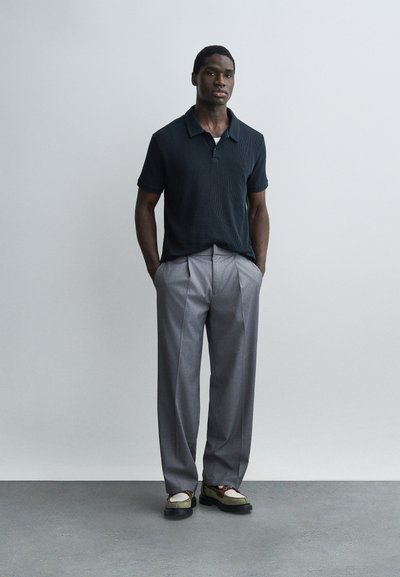 Homme debout les mains dans les poches, portant un polo noir, un pantalon plissé gris et des mocassins bicolores sur un sol gris.