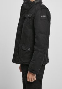 Muž v černé zapnuté bundě s klopovými kapsami stojí bokem proti jednobarevnému pozadí.