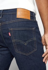 Jean Levi’s bleu foncé avec poche arrière visible, écusson en cuir de la marque et une main reposant près de la hanche, porté avec une chemise blanche.