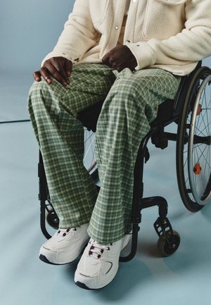 Pantaloni a quadri verdi con una vestibilità ampia, giacca in pile color crema e scarpe bianche con accenti neri, indossati da una persona in sedia a rotelle.