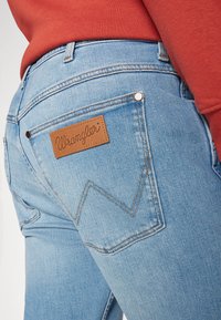 Svijetloplave traperice s kožnim Wrangler logom. Karakteristike uključuju cik-cak šav na stražnjem džepu i metalne gumbe kao dekoraciju.