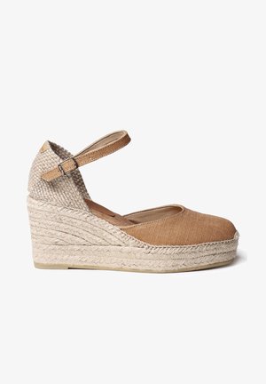 Zapato alpargata con cuña y plataforma de yute tejido, parte superior de lona color tan y correa ajustable en el tobillo con hebilla sobre fondo blanco.