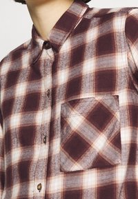 Karierter Flanellhemd in Burgunderrot, Grau und Creme, mit einem umlegbaren Kragen, vorderen Knöpfen und einer rechteckigen Brusttasche. Weiche Textur.