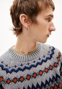 Grauer gestrickter Pullover mit einem geometrischen Muster in Blau, Orange und Schwarz. Hat einen gerippten Halsausschnitt und wird über einem beigen Hemd getragen.