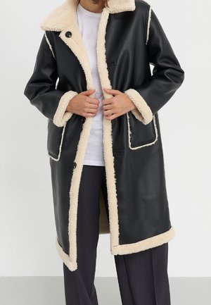 Person iført en lang sort læderfrakke med beige shearlingkant, hvid skjorte og mørke bukser, justerer fronten på frakken.