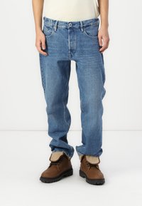 Blauwe spijkerbroek met een relaxte pasvorm, vijf zakken en een medium wash. Draagt met bruin gevlochten laarzen met een zachte voering bij de kraag.