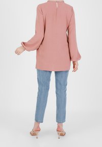 Bluzka z długim rękawem w odcieniu różowym, z dziurką na plecach, zestawiona z jasnoniebieskimi dżinsami o prostych nogawkach i cielistymi sandałami na obcasie. Gładka faktura materiału.