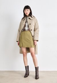 Beige gewatteerde jas met grote knopen, een geruite blouse, olijfgroene minirok met splits, en donkerbruine instappers met hak.