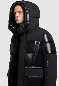 Must must jacket läikiva viimistlusega, üle mõõtmete kapuuts, lukuga tasku ja reguleeritavad nööbid. Kujunduses on kombineeritud mati ja läikiva tekstuuri.