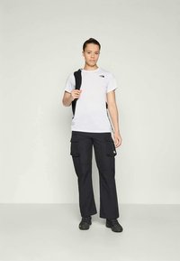 Donna con indosso una maglietta bianca North Face, pantaloni cargo neri, sneakers nere e una borsa a zainetto nera a tracolla su una spalla.