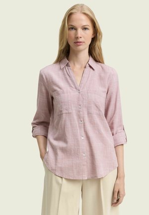 Donna che indossa una camicia rosa chiaro con bottoni e maniche rimboccate, con pantaloni beige, in piedi contro uno sfondo neutro con le mani nelle tasche.