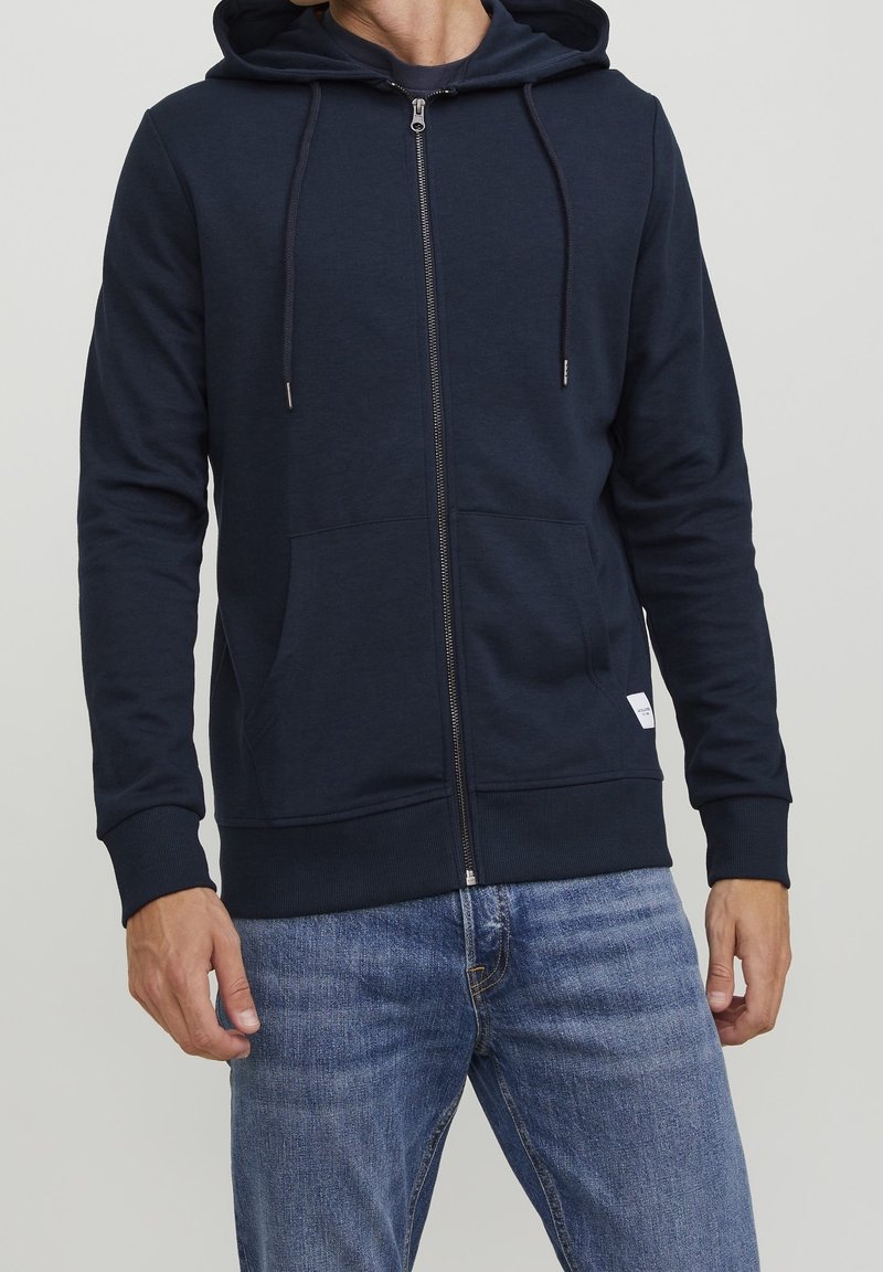 Homme portant un sweat à capuche zippé bleu marine avec poches avant et cordons, associé à un jean en denim bleu sur un fond clair.