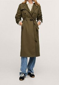 Olivenfarvet trenchcoat med bælte i taljen, dobbeltradet design og knapfastgjorte skulderepauletter, parret med løse blå jeans og sneakers.