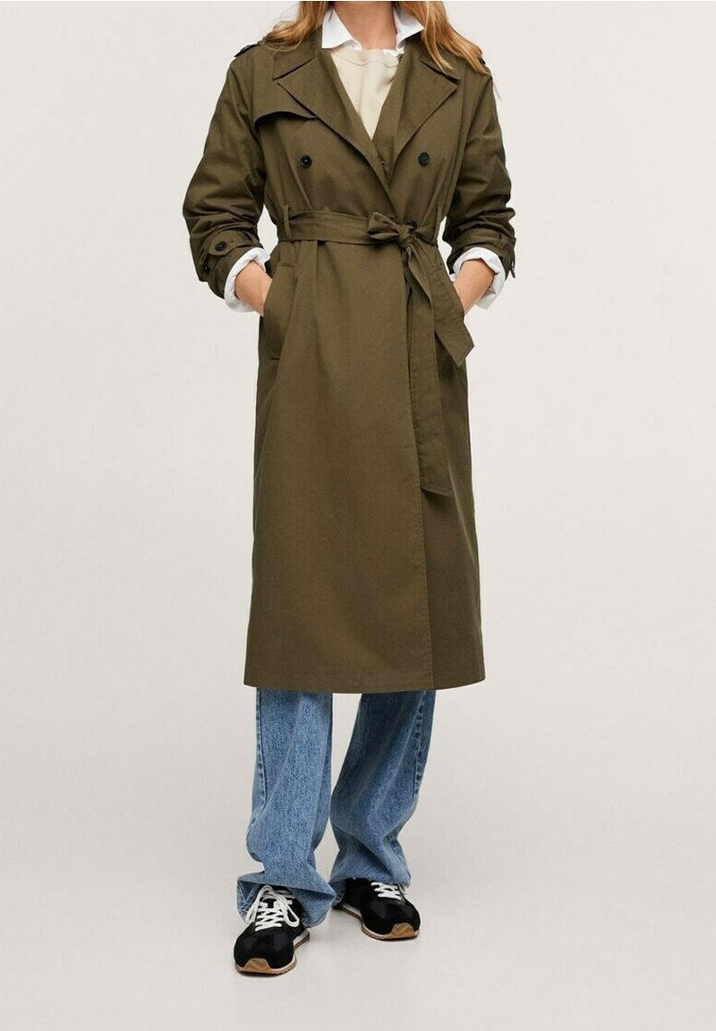 Olivenfarvet trenchcoat med bælte i taljen, dobbeltradet design og knapfastgjorte skulderepauletter, parret med løse blå jeans og sneakers.