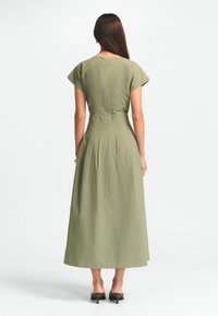 Oliivinvihreä mekko, jossa on lyhyet hihat, vartaloa myötäilevä yläosa ja virtaava, vartalonmyötäinen hame. Takana vetoketju, sileä kankaan rakenne. Korkokengät.