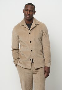 Beige corduroy jas met een notch kraag, knoopsluiting en ribbelstructuur. Draag het over een zwart shirt, gecombineerd met bijpassende broeken.