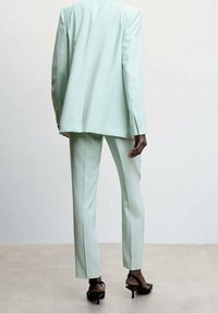 Personne portant un costume sur mesure vert menthe clair et des sandales à talons noires, debout devant un fond uni gris clair.