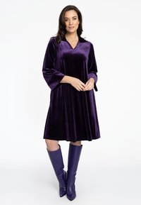 Lila Samtkleid mit V-Ausschnitt, Ärmeln bis zur Elle und ausgestellter Form. Getragen mit kniehohen lila Stiefeln, beide glatt und glänzend.