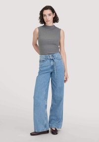 Graues, ärmelloses Oberteil, kombiniert mit hoch taillierten, weit geschnittenen, hellblauen Jeans, die einen gebundenen Bund und eine glatte Textur aufweisen, abgerundet mit dunklem Schuhwerk.