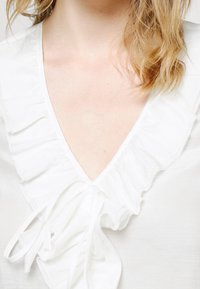 Blouse blanche à col en V profond, avec un devant à volants et un détail à nouer. Tissu doux et léger avec un léger éclat.
