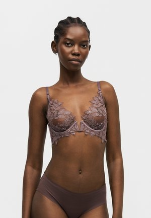 Kvinde iført en malva blonde-bh med bøjle og blomsterdesign samt matchende glatte hipster-trusser på ensartet lys baggrund.