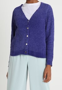 Fialový žebrovaný kardigan s V-výstřihem, předním zapínáním na knoflíky a dlouhými rukávy, vyrobený z měkkého pleteného materiálu. V kombinaci se světle modrými kalhotami.