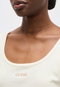 Κοντινό πλάνο ενός ατόμου που φοράει λευκό ριμπαριστό αμάνικο μπλουζάκι με το λογότυπο "GUESS" κεντημένο σε μπεζ χρώμα στο στήθος, εμφανίζοντας τον λαιμό και τους πάνω ώμους.