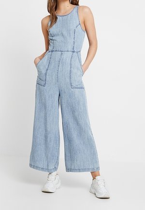 Kvinde iført ærmeløs lyseblå denim jumpsuit med vide ben og forlommer, kombineret med hvide sneakers.