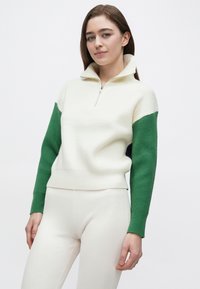 Ribstrikket halvlange lynlås-pullover i hvid med grønne ærmer, med høj krave og tætsiddende pasform. Kombineret med matchende lyse bukser.