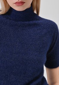 Donna che indossa un maglione scollato a collo alto a maniche corte di un blu scuro e peloso, con dettagli in filo metallico, su uno sfondo chiaro e semplice.