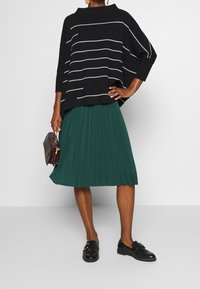 Schwarzes Strickoberteil mit weißen horizontalen Streifen, kombiniert mit einem grünen plissierten Rock und schwarzen Loafers. Modell hält eine dunkelbraune Handtasche.