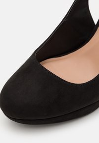 Chaussure à talon haut en suède noir avec un bout rond, un côté ouvert et une doublure intérieure lisse. La chaussure présente des détails de couture minimalistes.