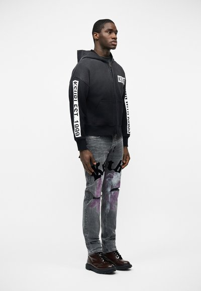 Άνδρας που φοράει μαύρο φούτερ με φερμουάρ και άσπρα γράμματα στα μανίκια και γκρι τζιν με μαύρα και μοβ γραφικά, στέκεται στραμμένος προς τα δεξιά.