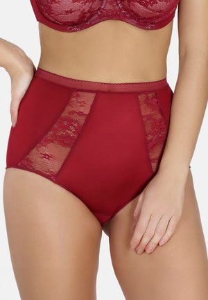 Woman wearing high-waisted red panties with floral lace panels and matching red lace bra, hand resting on hip.