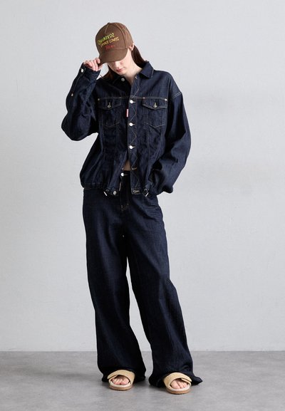 Denim jacket with two chest pockets, gold stitching, paired with loose-fitting dark denim pants and a brown cap featuring neon embroidery.