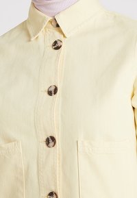 Chaqueta amarilla clara de botonadura con grandes botones marrones, dos bolsillos frontales, llevada sobre un cuello de tortuga de canalé rosa claro.