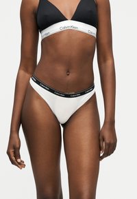 Γυναίκα που φοράει μαύρο και λευκό σουτιέν Calvin Klein και λευκά εσώρουχα Calvin Klein με μαύρη ζώνη, στέκεται πάνω σε απλό φόντο.