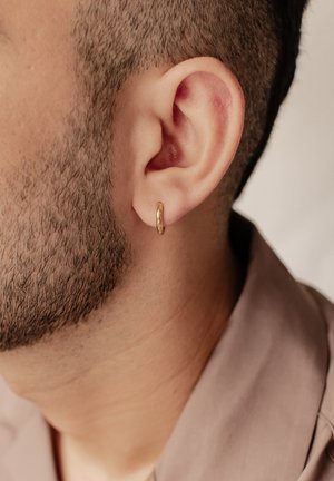 Pendiente de aro dorado en una oreja con barba y pelo corto, que presenta un acabado liso y pulido y un diseño circular.