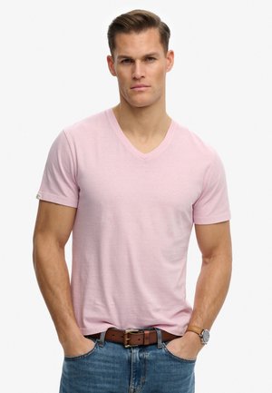 Uomo con capelli castani corti indossa una T-shirt a V rosa chiaro, jeans blu, cintura marrone e orologio da polso, in piedi con le mani nelle tasche.