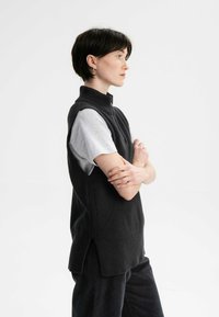 Schwarzer, gestrickter ärmelloser Rollkragenpullover mit seitlichem Schlitz, über einem hellgrauen Kurzarmshirt getragen. Auf der gesamten Fläche mit einem strukturierten Muster versehen.