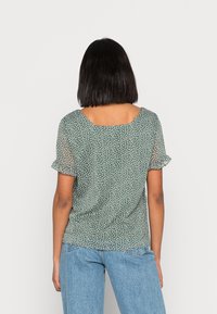 Kvinna med rakt svart hår som bär en grön kortärmad blus med vita prickar och ljusblå jeans, sedd bakifrån.