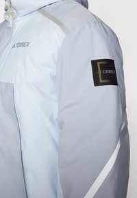 Veste bleu clair avec capuche, présentant un écusson noir avec un logo, des accents argentés sur les épaules et une texture lisse et mate.