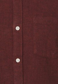 Bordeaux farbener Button-Up-Hemd aus strukturiertem Stoff, mit einer Brusttasche und vier weißen Knöpfen an der Vorderseite.