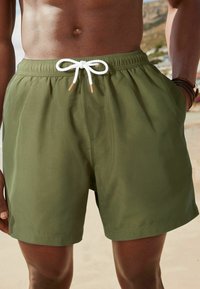 Zöld úszónadrág könnyű anyagból, elasztikus derékkal, fehér húzózsinórral és bézs végződéssel. Simább textúra, térd hosszúságú.